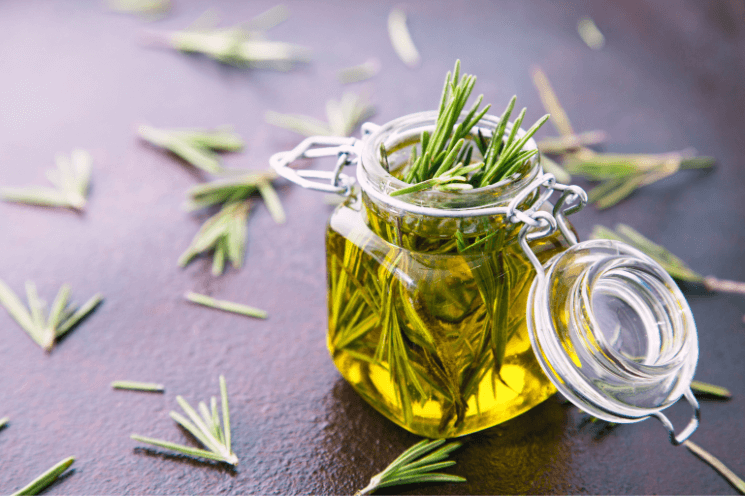 jar of rosemary oil, with fresh rosemary leaves scattered around