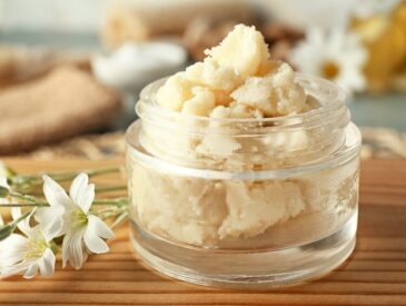 glass jar of shea butter on wooden surface with small white flowers beside it